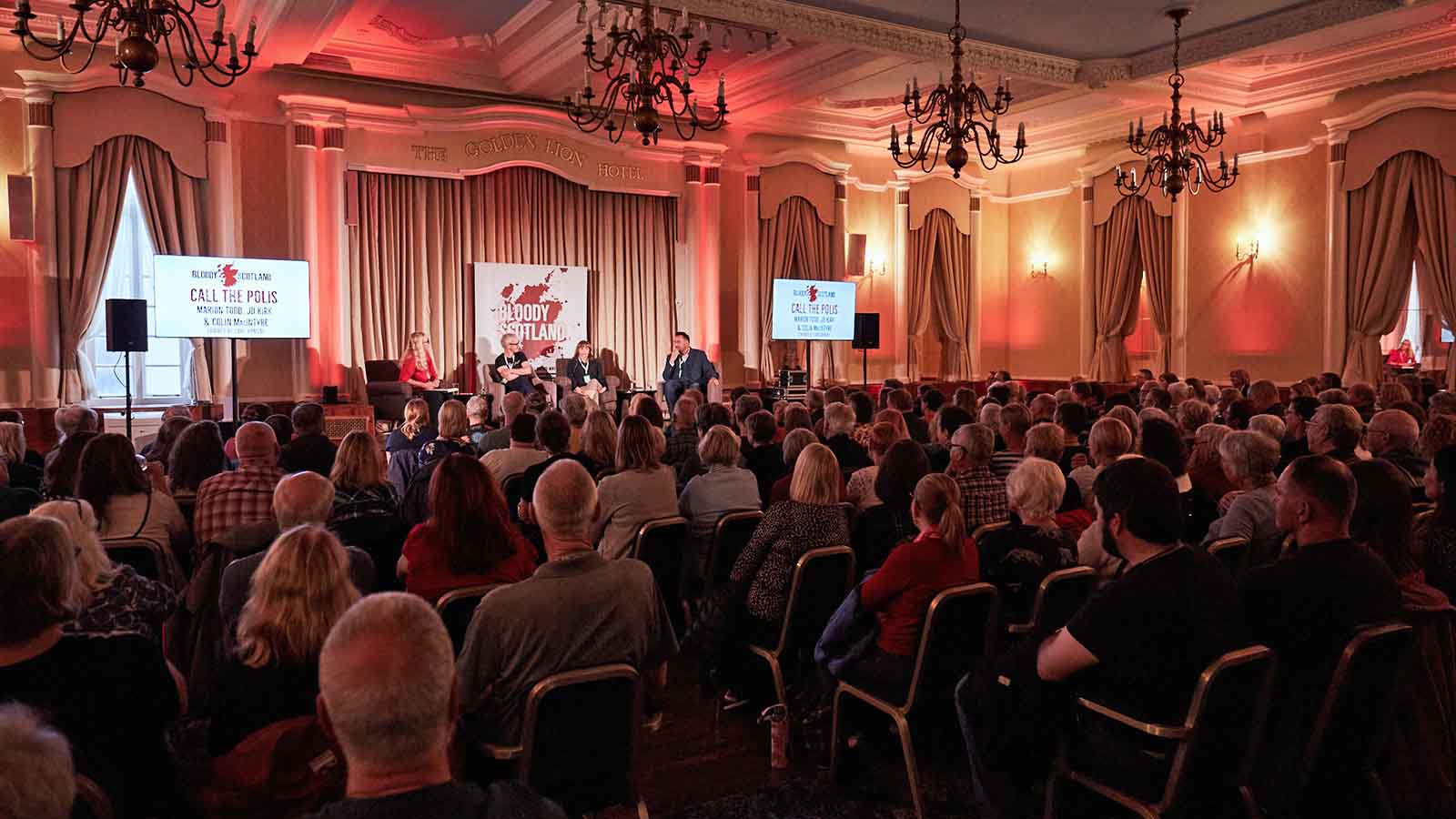 A large dimly lit room and a big audience of people seated watching speakers on a stage.