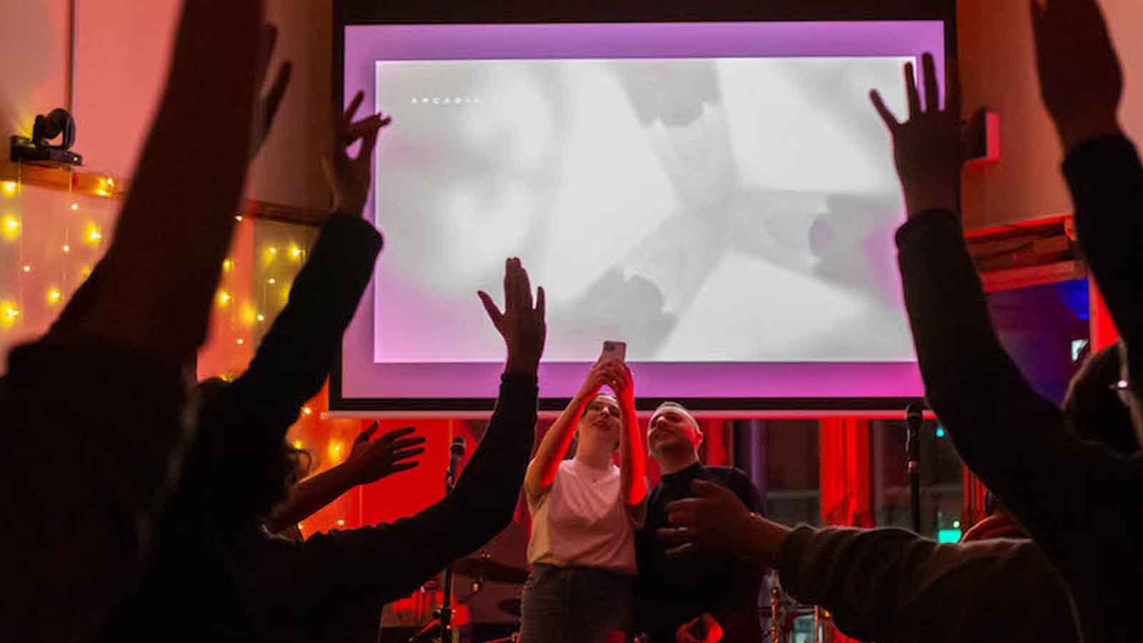 In a darkened room lit by fairy lights and red lighting, two people stand in front of a projected screen taking a phone photo of the crowd who have their hands raised in the air.