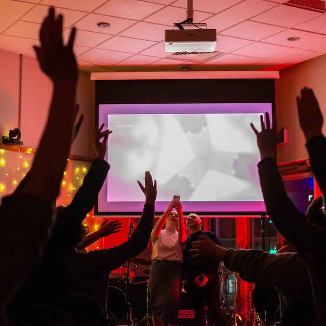 In a darkened room lit by fairy lights and red lighting, two people stand in front of a projected screen taking a phone photo of the crowd who have their hands raised in the air.