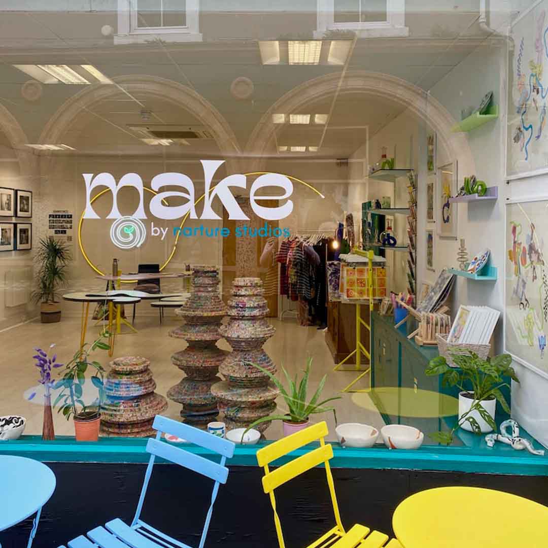 Exterior photo of a shop – the window vinyl reads 'Make by narture studios', above brightly coloured outdoor tables and chairs. Through the window is a bright white space with framed artworks on the walls and prints and sculpture placed around the room.