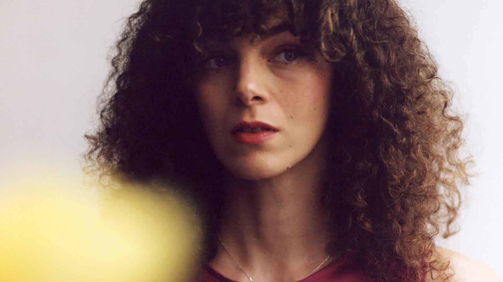 A woman with lots of dark curly brown hair, her face partially lit by a bright yellow blurred light in the corner