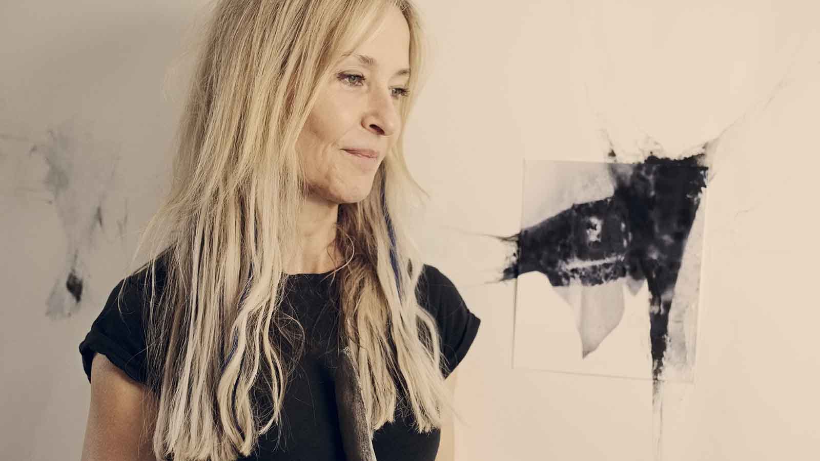 A woman with long blonde hair wearing a black top looking pensive. In the background black ink-like blotches on paper and the walls form abstract shapes.