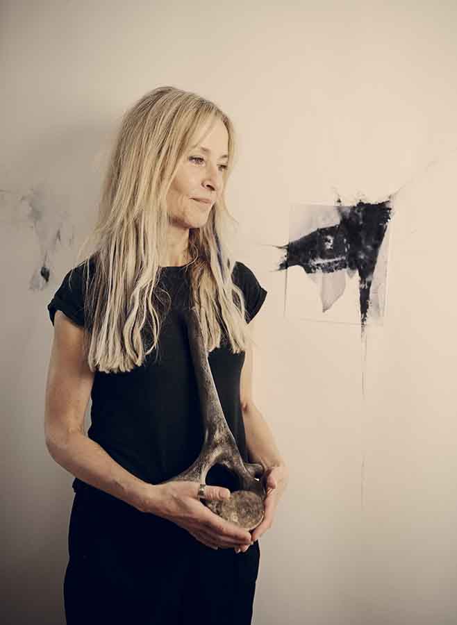 A woman with long blonde hair wearing a black top looking pensive. In the background black ink-like blotches on paper and the walls form abstract shapes.