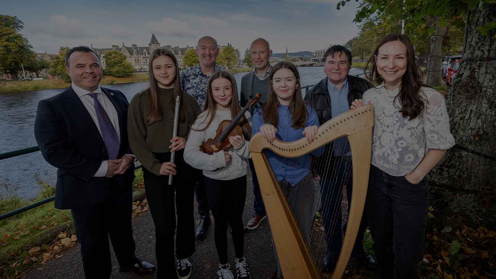 Eight people posing together on the banks of a river which runs in the background. Three of them are young women, holding musical instruments (a flute, a fiddle and a harp).