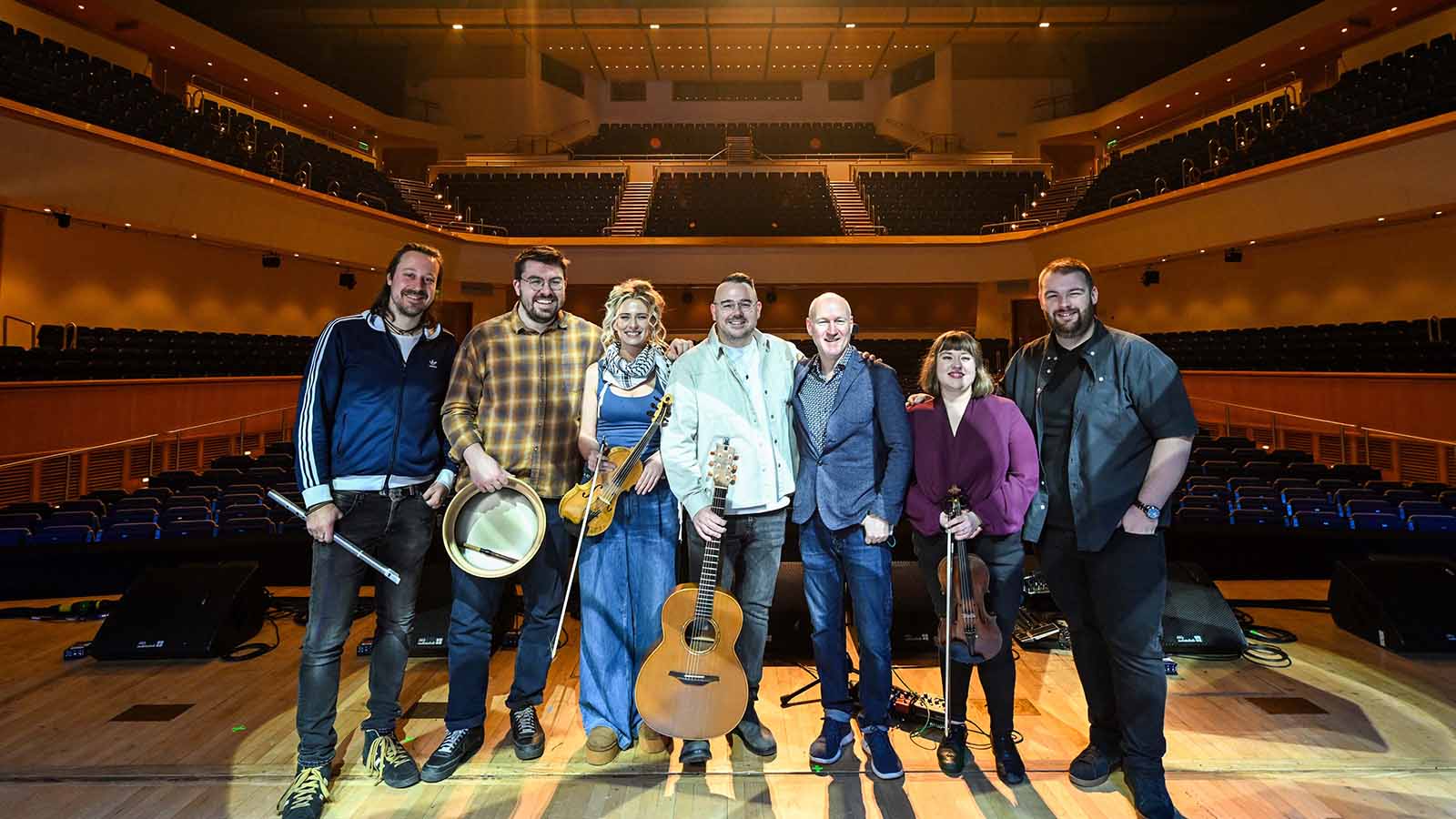 A group of musicians holding their instruments and Celtic Connections event organisers (Ross Ainslie, Craig Baxter, Aileen Reid, Paul McKenna, Donald Shaw, Madeline Stewart and Rory Matheson) stand on a stage with a large auditorium in the background.