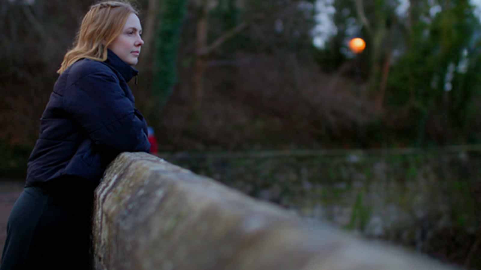 A woman with light red hair looks out as she leans on the wall of a stone bridge on a bright, cold day