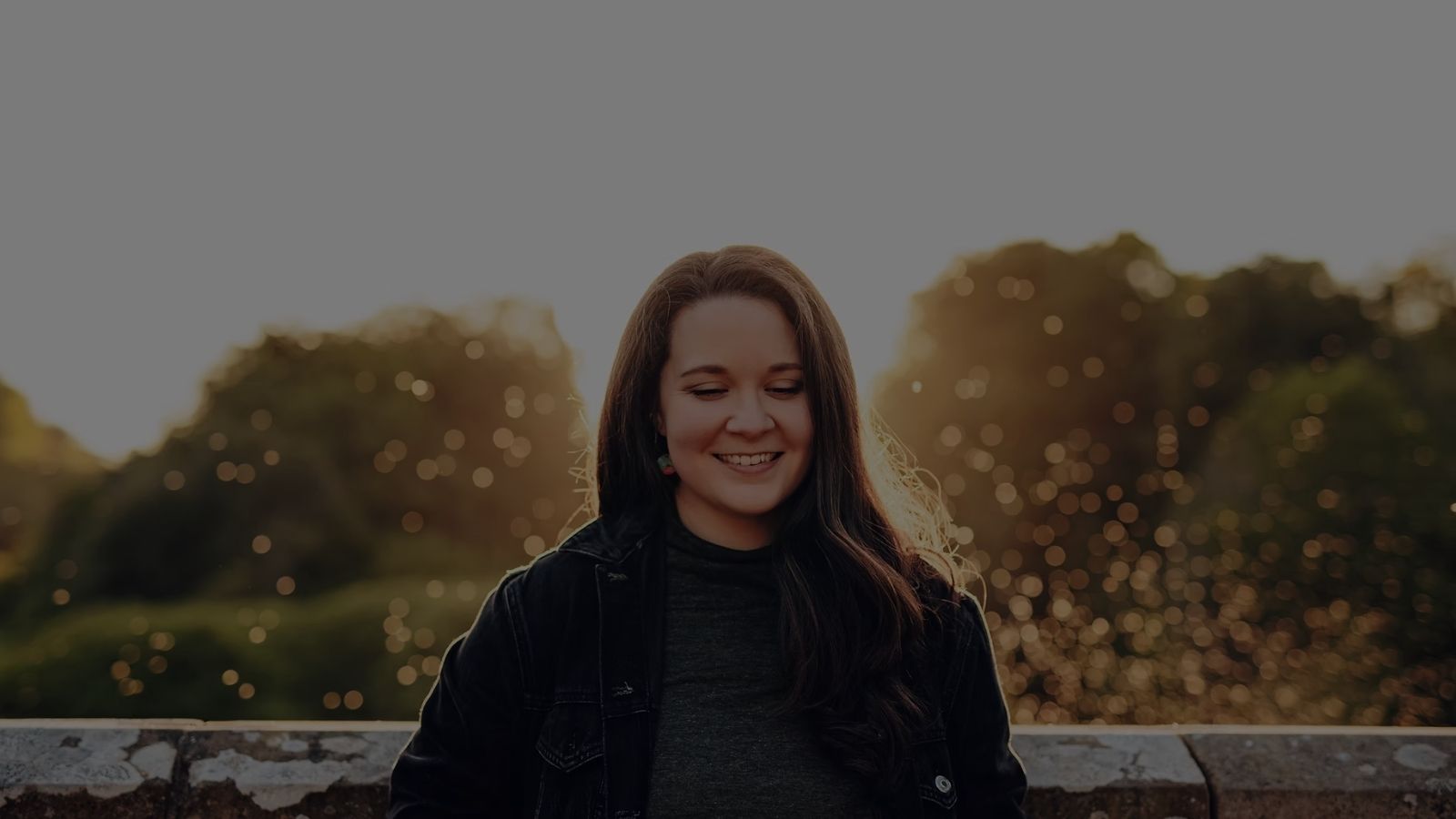 A woman with long dark brown hair silhouetted by a setting sun on a clear day, behind her is a low stone wall, this is Rachel Lightbody.