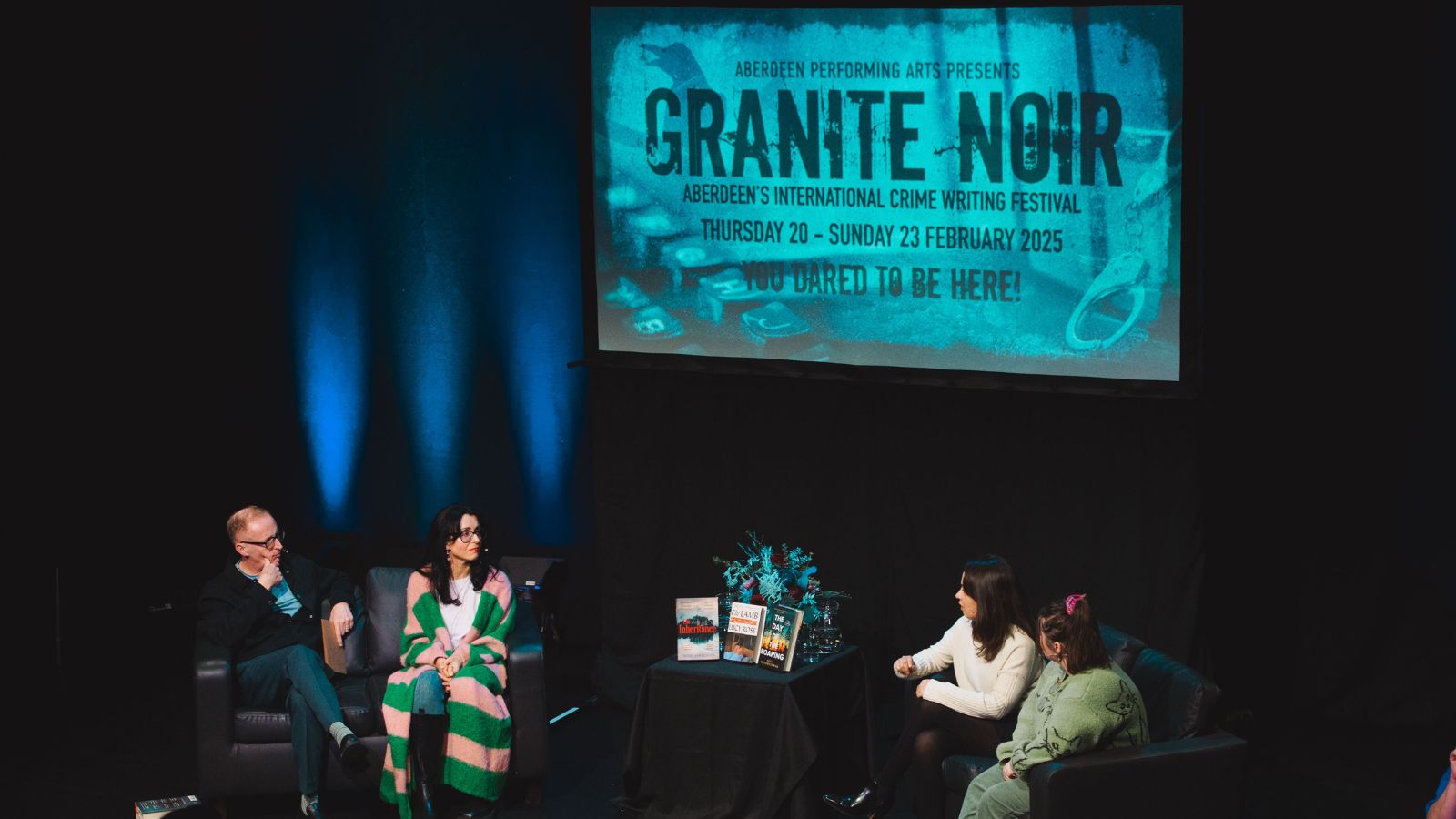 Four people sitting on large armchairs on a stage talking to each other. Above them in a large blue projection of the Granite Noir logo.