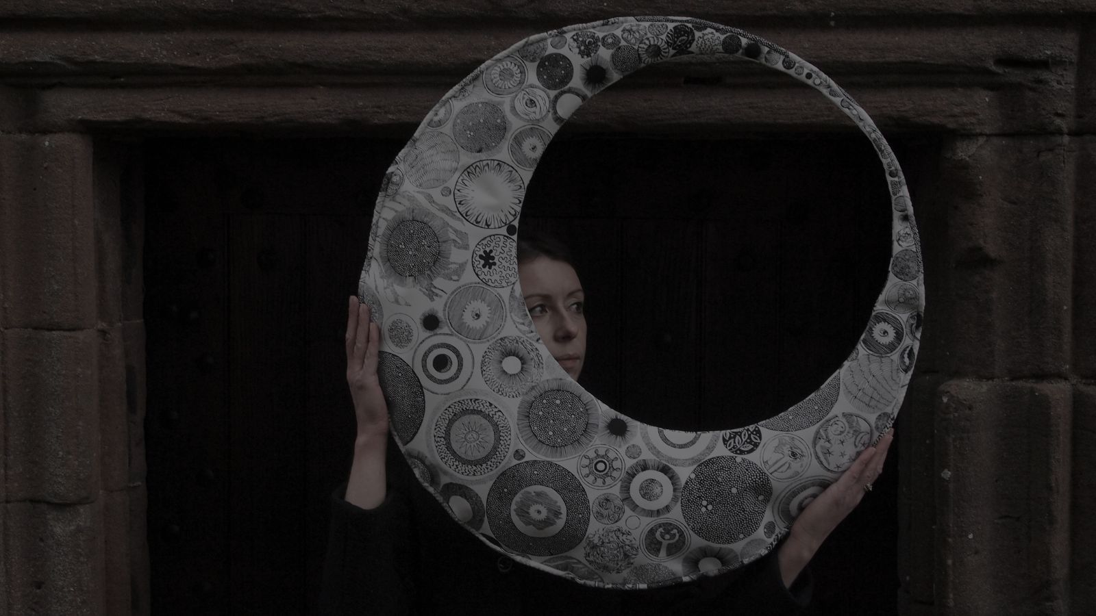 A woman with long brown hair wearing a black top and holding an intricately patterned object shaped like a closed crescent moon in front of a stone doorway. This is visual artist Helen Gallogly.