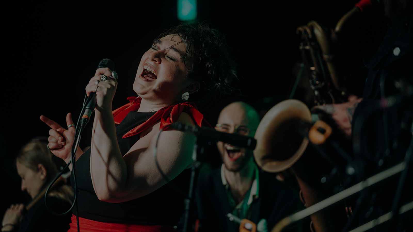 A woman in a black and red dress sings passionately into a microphone. In the background, out of focus a band member smiles excitedly as he watches her.