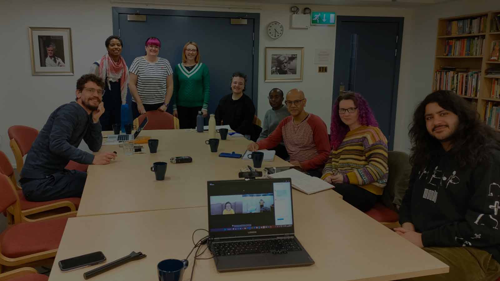 A group of adults posing together, some seated and some standing around a large wooden table covered in coffee cups and notes and pen, in a well-lit room