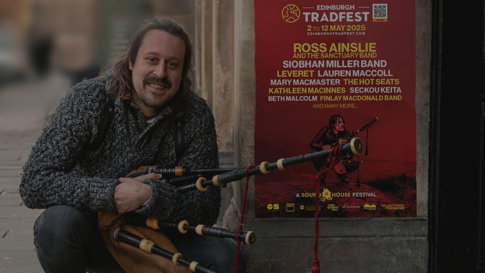 A man with shoulder-length brown hair and a beard, wearing a grey knitted jumper, crouches on a city street while holding a set of bagpipes. He is smiling at the camera. Behind him, a red poster for Edinburgh Tradfest 2025 is displayed on a stone wall. The poster features a list of performing artists, including Ross Ainslie and the Sanctuary Band, Siobhan Miller Band, and others. The event dates, 2–12 May 2025, are prominently shown, along with sponsor logos at the bottom