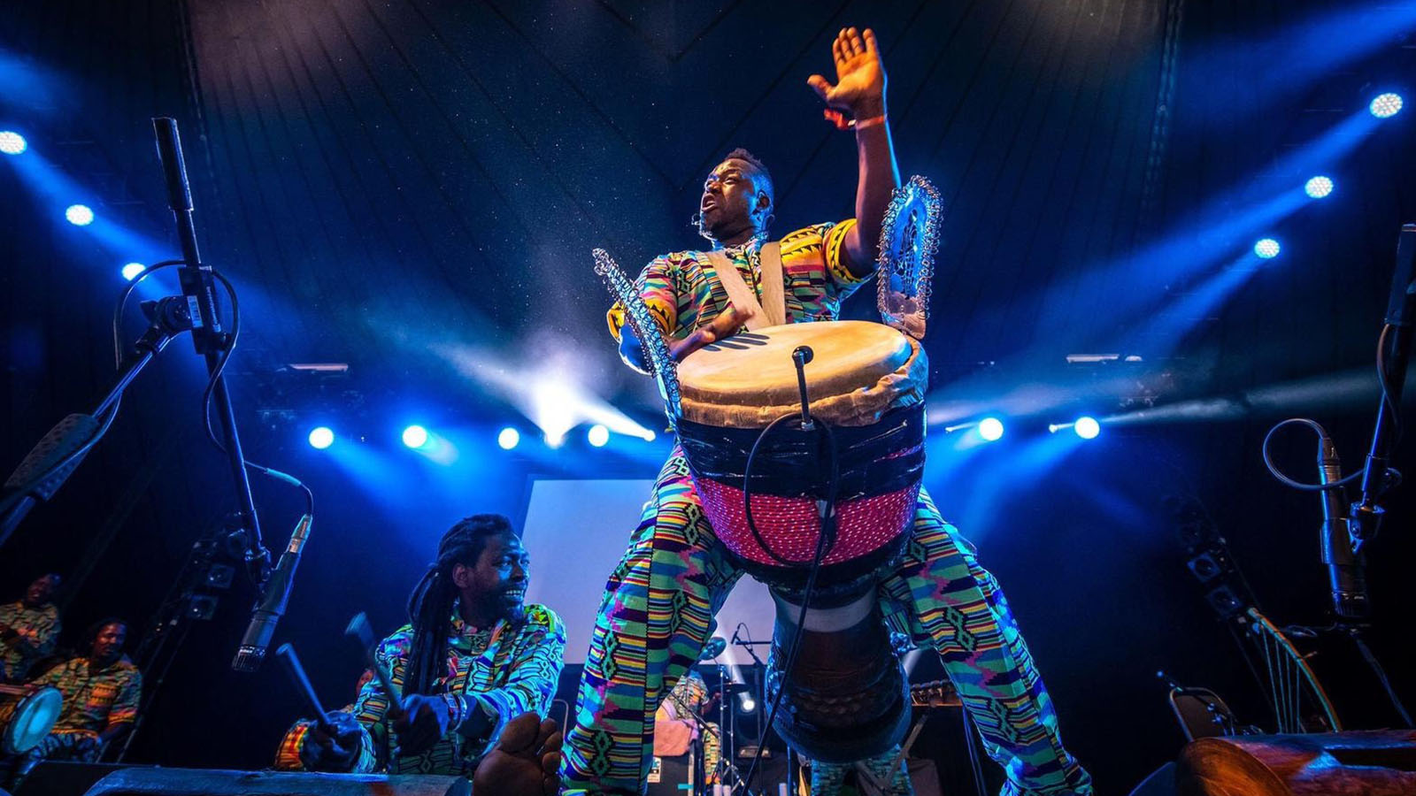 A dynamic stage performance featuring a djembe drummer in a vibrant, patterned outfit, captured mid-motion with one hand raised and the other striking the drum. Blue stage lighting and beams of light create a dramatic atmosphere. Other musicians in matching attire play percussion instruments in the background.