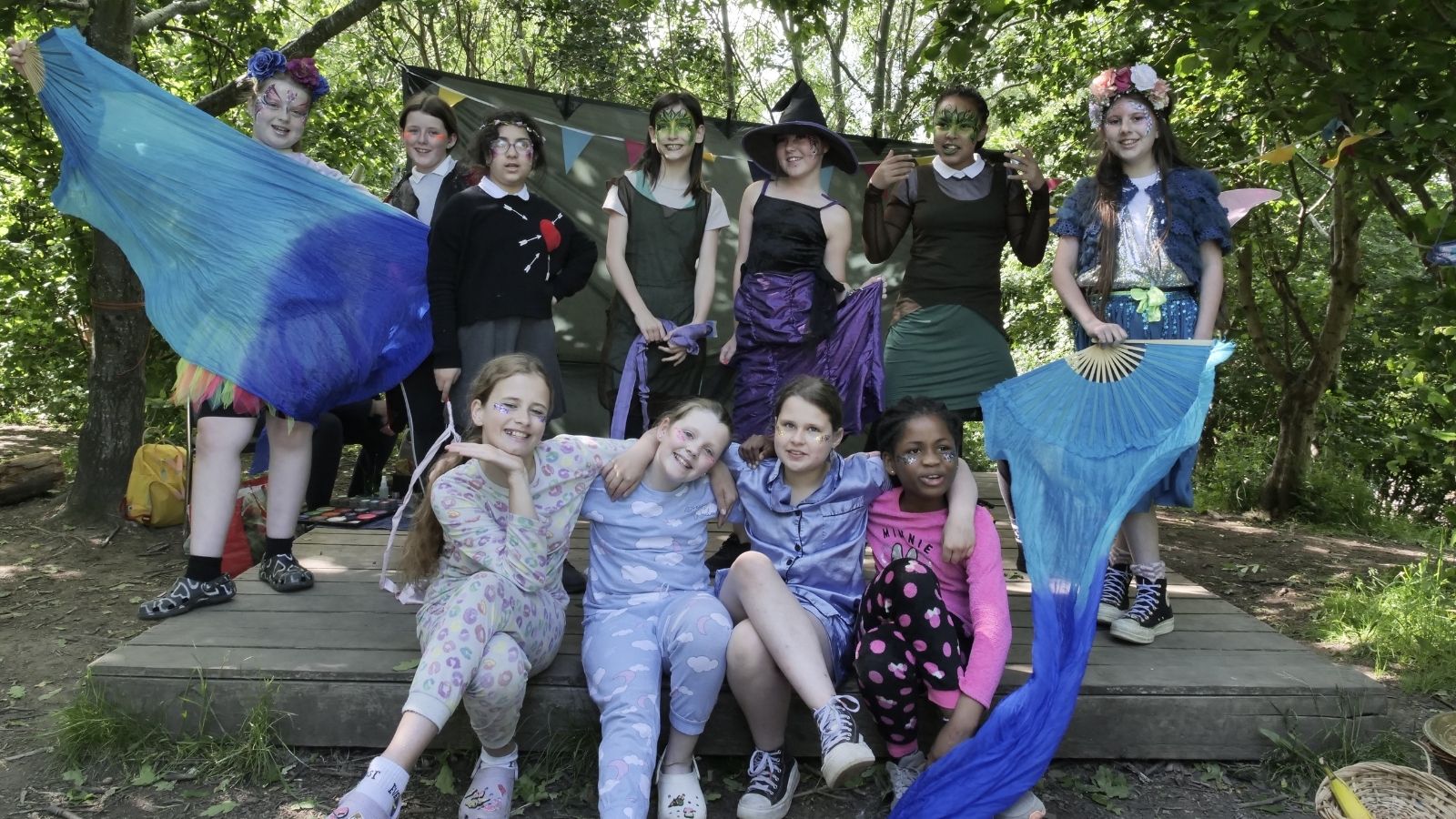 A large group of children and young people posing on an outdoor stage in the woods, on a sunny day