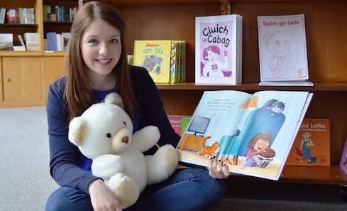 A woman sits on the floor in a library in front of Gaelic books, holding a stuffed toy