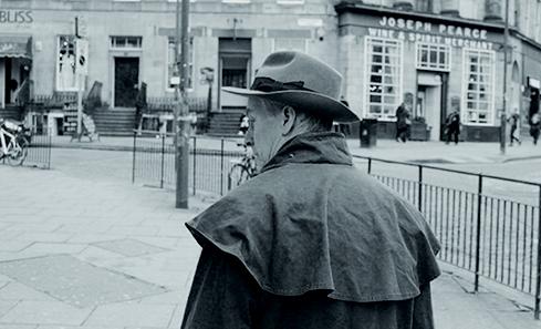A black and white image of a man wearing a hat walking through the city centre of Edinburgh - the bar Joseph Pearce is visible in the distance